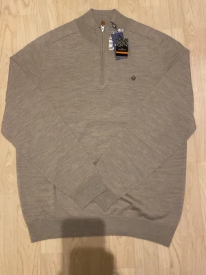  Morris - Snygg beige tröja från Morris i mjuk merinoull. Tröjan har half zip-dragkedja, ribbade muddar och en liten broderad logga på bröstet. Perfekt för dig som gillar stilrena och klassiska plagg med modern touch.