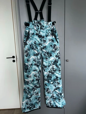 Blommiga täckbyxor från Everest - Säljer ett par blåa täckbyxor från Everest i storlek 164. Byxorna har dragkedjor vid fickorna och slitsar vid bensluten som går att stänga/öppna. Går att dra åt i midjan och går även att justera/ta bort hängslen.