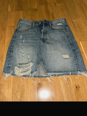 Blå jeanskjol med slitningar från Hm - Säljer en blå jeanskjol från HM med rå kant och slitna detaljer framtill och baktill. Kjolen har klassisk femficksdesign, knappar i gylfen och en rak passform. Perfekt för dig som gillar en avslappnad och trendig look.