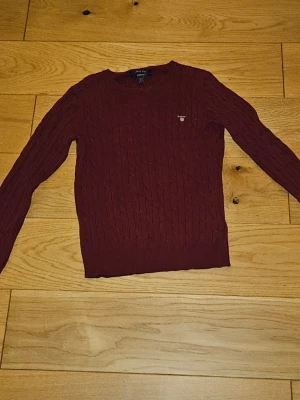 Vinröd kabelstickad tröja från GANT - Säljer en vinröd kabelstickad tröja från GANT i stretchig bomull. Tröjan har rund hals, diskret GANT-logo på bröstet och långa ärmar. Perfekt till höst och vinter för en klassisk och clean look. Värt att nämna är att den är liten i storlek så passar folk med S