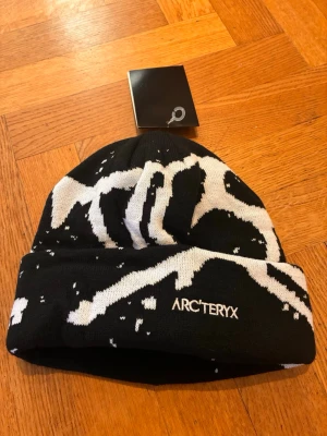 Svart och vit mössa från Arc'teryx - Säljer en svart och vit mössa från Arc'teryx med coolt abstrakt mönster. Mössan är stickad i mjukt material och har uppvikt kant med broderad logga framtill. Perfekt för kalla dagar och snygg till streetwear-stilen.