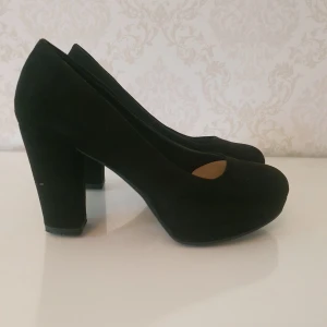 Svarta pumps med hög klack - Svarta pumps med rund tå och hög klack i storlek 37. Skorna har en plattform framtill och är klädda i mockaliknande material. Pumpsen är i det stora hela i fint skick och har ytterst små defekter på klacken och framtill. 