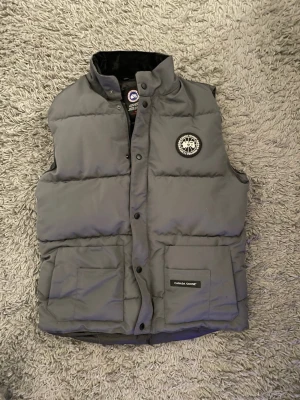 Grå dunväst från Canada Goose - Säljer en grå dunväst från Canada Goose med klassisk logotyp på bröstet. Västen har hög krage, tryckknappar framtill och två stora fickor med lock. Perfekt för kyliga dagar och har en stilren, quiltad design.