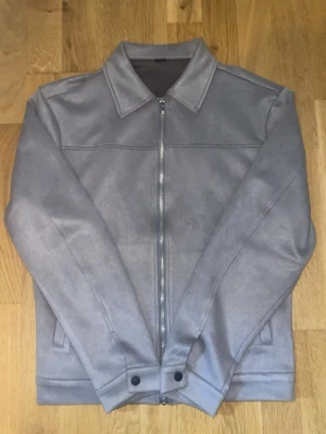 Grå mockajacka med dragkedja - Säljer en stilren grå mockajacka med klassisk krage och hel dragkedja framtill. Jackan har fickor på utsidan samt insidan. Jackan har raka linjer, diskreta sidfickor och knappdetaljer vid ärmsluten. Perfekt för dig som gillar minimalistisk och clean stil. Materialet är påriktigt wow, tack mvh 😊.