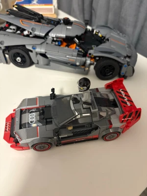 Grå och röd LEGO Audi e-tron väst - Säljer en LEGO-minifigur med en grå väst med Audi-logga på ryggen. Västen har sportig look, raka linjer och detaljer i svart och vitt. Perfekt för dig som gillar racing och coola bilar. Passar till minifigurer och är ett måste för alla bilnördar.