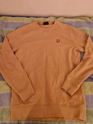 Beige sweatshirt från Lyle & Scott - Säljer en beige sweatshirt från Lyle & Scott med klassisk rund hals och ribbade muddar. Tröjan har det ikoniska gula fågelmärket broderat på bröstet och är tillverkad i mjuk bomull. Perfekt för dig som gillar enkel och stilren design.
