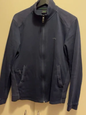 Marinblå ziptröja från BOSS - Säljer en marinblå ziptröja från BOSS med hög krage och diskret logga på bröstet. Tröjan har två fickor med dragkedja och är tillverkad i mjuk bomull. Perfekt för lager på lager och chill vardagsstil.