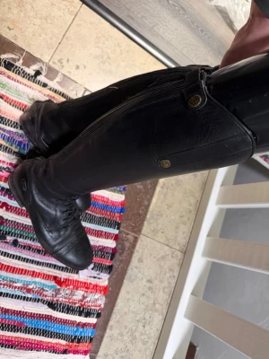 Svarta ridstövlar i skinn - Säljer ett par höga svarta ridstövlar i skinn med snörning framtill och dragkedja där bak. Stövlarna har dekorativa detaljer och knappar upptill samt ribbade partier för extra flexibilitet. Perfekta för dig som gillar klassisk ridstil.