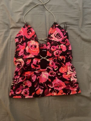 Blommig halterneck topp från Bik Bok - Supersnygg blommig halterneck topp från Bik Bok i starka rosa, röda och svarta toner. Toppen har glittriga snörningsdetaljer framtill och runt halsen, samt en djup v-ringning. Perfekt för dig som vill sticka ut med färg och detaljer.