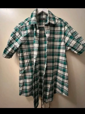 Grön och vit rutig skjorta kort ärmad - Helt ny, oanvänd kortärmad skjorta i grön och vit rutig design. Skjortan har klassisk krage, knäppning framtill och perfekt för dig som gillar en avslappnad men ändå stilren look.