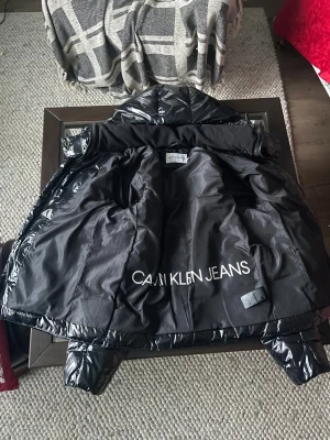 Svart glansjacka från Calvin Klein - Säljer en svart, glansig pufferjacka från Calvin Klein Jeans med huva och dragkedja. Jackan har en quiltad design, logga på bröstet och stor Calvin Klein Jeans-tryck på insidan. Perfekt för kalla dagar och riktigt snygg streetstil. Kan diskutera pris.