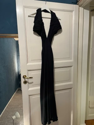 Svart långklänning med slits - Supersnygg svart långklänning med djup v-ringning och markerade axlar. Klänningen har en hög slits framtill och är figurnära upptill med draperade detaljer. Perfekt för bal eller fest. Har en liten söm som gått up vid axeln 