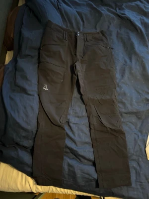 Svarta täckbyxor från Haglöfs herr - Säljer ett par svarta täckbyxor från Haglöfs för herr. Byxorna har dragkedjor på fickorna, förstärkta knän och justerbara benslut med kardborre. Perfekta för utomhusaktiviteter och äventyr. Materialet är slitstarkt och designen är funktionell.