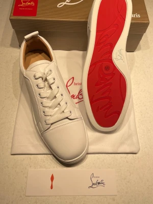 Vita sneakers från Christian Louboutin - Säljer ett par stilrena vita sneakers från Christian Louboutin i skinn med klassisk röd sula. Skorna har rund tå, snörning och diskret branding på sulan. Perfekta för dig som vill ha soprena och lyxiga sneakers med ikonisk design.