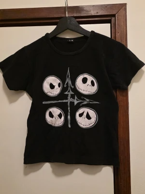 Nightmare Before Christmas t-shirt - Svart t-shirt med tryck av fyra olika Jack Skellington-ansikten på framsidan och stor text 'The Nightmare Before Christmas' på ryggen. Klassisk rund hals och korta ärmar. Perfekt för dig som älskar Tim Burton och vill ha en unik tee med ikoniskt motiv. Tröjan är mer Small än Medium.