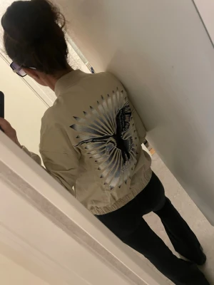 Beige bomberjacka med broderad fågel - Säljer en beige bomberjacka med stor broderad svart och silverfärgad fågel på ryggen. Jackan har dragkedja framtill, två stora fickor och ribbade muddar vid ärmar och midja. Materialet känns lätt och passar perfekt till streetwear.