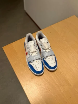 Nike Jordan 1 Low vit/blå/röd - Säljer ett par Nike Jordan 1 Low sneakers i vitt, blått och rött med gulddetaljer. Skorna har klassisk snörning, bred kardborreband med Nike-logga och perforerad tå. Swoosh i rött på sidan och Jumpman-logga i guld på tungan och hälen. Tillverkade i skinn och syntet.