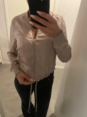 Beige bomberjacka med snörning - Säljer en beige bomberjacka med dragkedja framtill och snörning i midjan. Jackan har ribbade muddar vid ärmslut och hals, samt en stilren och enkel design. Perfekt för dig som gillar en avslappnad men snygg look.
