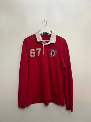 Röd långärmad vintage från Polo Ralph Lauren - Röd långärmad vintage från Polo Ralph Lauren med vit krage, stora siffror '67' på bröstet och ryggen samt patch med amerikansk flagga på ryggen. Skjortan har även ett broderat märke på bröstet och knappar vid halsen.
