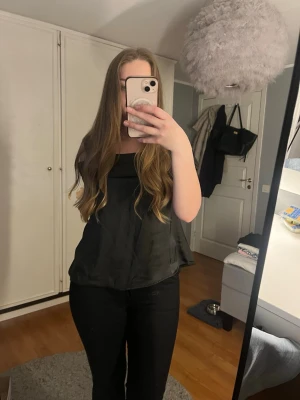 Svart omlott topp från NA-KD - Snygg svart omlott topp från NA-KD i glansigt satintyg. Toppen har breda axelband, offshoulder-detalj och en stor rosett i ryggen. Modellen är ärmlös och har en öppen rygg som ger en trendig look. Perfekt att styla med jeans eller kjol. Aldrig blivit använd, köptes för ca 400kr.