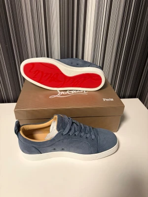 Blå Christian Louboutin sneakers - Snygga blå sneakers från Christian Louboutin med klassisk röd sula och vit kant. Skorna är gjorda i mocka och har snörning samt rund tå. Insidan är beige och ger en lyxig känsla. Perfekta för dig som vill sticka ut med exklusiva detaljer. Storlek 43, helt nya och box finns!