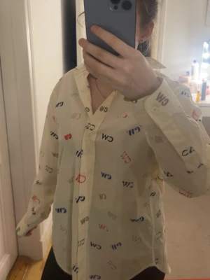 Vit skjorta med CW-print från C.W. - Säljer en vit skjorta från C.W. i storlek 32. Skjortan har ett färgglatt all-over CW-print i blått, rött, beige och grått. Klassisk krage, lång ärm och knäppning framtill. Tillverkad i tunn bomull som känns lätt och skön.