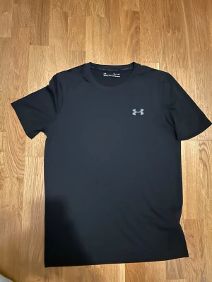 Svart t-shirt från Under Armour - Säljer en svart t-shirt från Under Armour med klassisk logga på bröstet. T-shirten är kortärmad och gjord i ett mjukt och lätt syntetmaterial som andas, perfekt för träning eller chill. Enkel och stilren design som funkar till det mesta.
