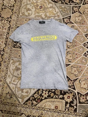 Grå DSQUARED2 t-shirt med tryck - Snygg grå t-shirt från DSQUARED2 med gul logga tryckt på bröstet. Klassisk passform och rund hals. Tillverkad i mjuk bomull som känns skön mot huden. Perfekt för dig som gillar streetwear och vill ha något stilrent men ändå med edge.