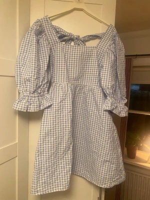 Blåvit rutig kortklänning med puffärm - Supersöt blåvit rutig kortklänning med puffiga långärmar och volang vid ärmslut som ger lite Alice i underlandet vibes. Klänningen har fickor, knyt i ryggen och dragkedja i sidan. Perfekt till sommaren!🌸