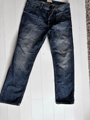 Mörkblå jeans från Jack & Jones - Säljer ett par mörkblå jeans från Jack & Jones med klassisk femficksdesign och raka ben. Jeansen har en snygg tvätt med lätt slitning och knappar i gylfen. Perfekta för dig som gillar en enkel och tidlös stil. Skick 9/10