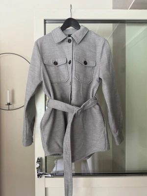 Grå overshirt med bälte - Snygg grå overshirt med knappar framtill, två stora bröstfickor och ett matchande bälte i midjan. Jackan har lång ärm och en klassisk krage. Perfekt för lager på lager och enkel att styla till olika outfits.