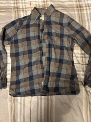 Rutig skjorta från Ruffle & Bow - Säljer en rutig skjorta från Ruffle & Bow i brunt, blått och grått. Skjortan har klassisk krage, knappar framtill och en bröstficka. Tillverkad i mjuk bomull och har långa ärmar. Perfekt för en avslappnad och cool stil.