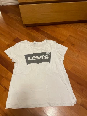 Vit Levi's t-shirt med logga - Vit t-shirt från Levi's med klassisk grå logga tryckt på bröstet. T-shirten har rund halsringning och korta ärmar. Tillverkad i mjuk bomull som känns skön mot huden. Perfekt till jeans eller shorts för en enkel och clean look.