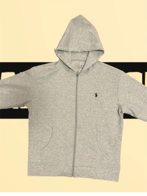 Grå Ralphlauren zip hoodie - Stilren och grisch huvtröja från Polo Ralph Lauren i ljusgrått. Dragkedja framtill, två fickor och klassisk broderad logga på bröstet. Mjuk bomullsblandning, normal passform. Mycket fint skick utan synliga slitage. Perfekt för både vardag och avslappnade tillfällen.