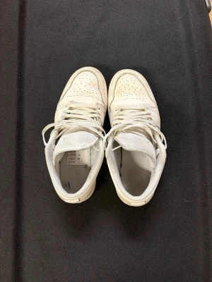 Nike Air Jordan 1 Low vita sneakers - Säljer ett par vita Nike Air Jordan 1 Low sneakers i skinn med klassisk broderad Air Jordan-logga på hälen och perforerad tå. Skorna har snörning och en platt sula. Perfekta för dig som gillar stilrena och ikoniska sneakers.