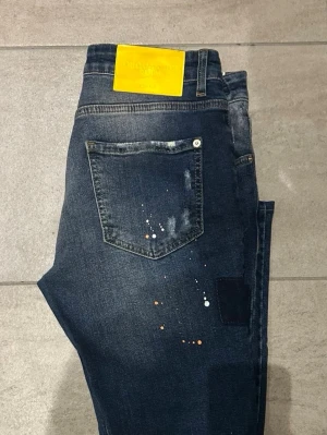 Dsquared2 blå jeans med färgstänk - Säljer ett par blå jeans från Dsquared2 med unika färgstänk i vitt och orange samt slitna detaljer och patchar på benen. Jeansen har klassisk femficksdesign, rak passform och en gul patch bak i midjan. Tillverkade i tåligt denimtyg.