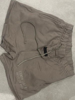 Beige Fear of God Essentials shorts - Säljer ett par beige shorts från Fear of God Essentials med långa snören i midjan och tryckt logga på benet. De har elastisk midja och är gjorda i mjuk bomull, perfekta för chill dagar. Klassisk loose fit och diskret färg som passar till allt.