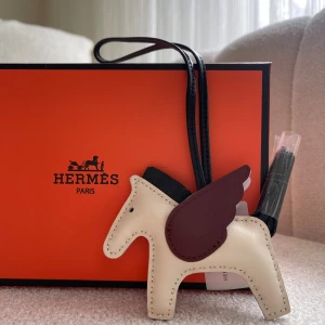  Pegasus väsktillbehör i skinn - Supersöt Pegasus-väsktillbehör från  i beige skinn med vinröda vingar och svart man. Perfekt som dekoration till din väska eller nyckelring. Lyxig och unik design med detaljerad sömnad och svart rem.