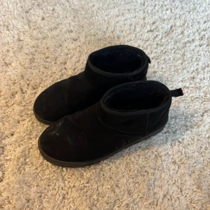 Svarta mockaboots med platt sula - Svarta boots i mocka med platt sula och rund tå. Skorna har en enkel design med låg skaft och enkel påtagning. Yttersulan är mönstrad för extra grepp. Perfekt för dig som gillar stilrena och bekväma skor. Liknande Uggs. Storlek 41 men lite små i storleken. De är lite smutsiga på bilderna, annars fint skick! 