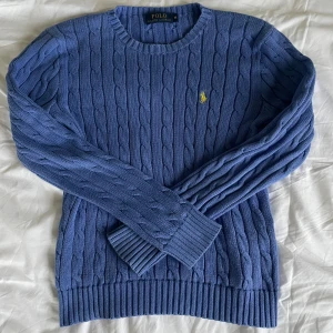 Blå kabelstickad tröja Polo Ralph Lauren - Kabelstickad blå tröja från Polo Ralph Lauren med rund halsringning och klassisk gul broderad logga på bröstet. Tröjan har ribbade muddar och är tillverkad i mjuk bomull. Storlek M men passar även en S