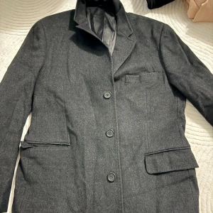 Mörkgrå kappa med tre knappar - Snygg mörkgrå kappa med klassisk passform och tre knappar framtill. Kavajen har två fickor med lock och en bröstficka. Perfekt för dig som vill ha en stilren och tidlös look. Fodrad insida för extra komfort.