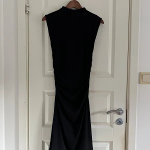 Svart långklänning från H&M - Svart långklänning från H&M i storlek S. Klänningen har ärmlös design, hög hals och snygg drapering längs sidorna som ger en cool siluett. Tillverkad i mjukt och stretchigt material som sitter bekvämt och följer kroppen.