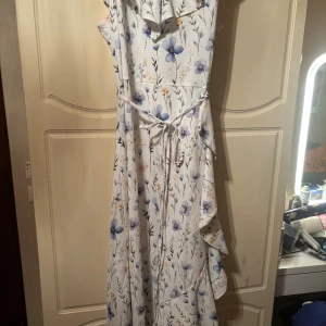 Blommig långklänning med volang - Superfin långklänning med blommigt mönster i lila, blå och gula toner på vit botten. Klänningen har volangdetaljer upptill och ett knytband i midjan för extra passform. Perfekt för sommarens alla tillfällen och har en luftig och skön känsla.