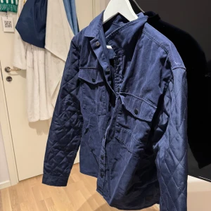 Mörkblå quiltad jacka Jack & Jones - Quiltad mörkblå jacka från Jack & Jones Core med två stora bröstfickor och knappar framtill. Jackan har långärmade ärmar med snyggt rutmönster på sidorna och är tillverkad i bomull. Perfekt för dig som gillar stilren och enkel design.