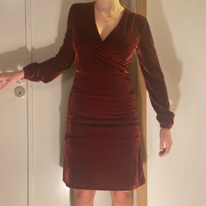 Vinröd omlottklänning från Indiska  - Supersnygg vinröd kortklänning från Indiska i mjuk sammet. Klänningen har omlottdesign med v-ringning, långa ärmar med lätt puff och markerad midja. Perfekt för dig som gillar elegant stil och vill sticka ut med en djup färg.