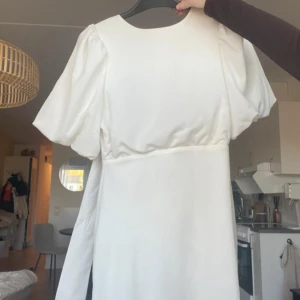 Vit kortklänning med puffärmar student  - Supersnygg vit kortklänning med puffiga kortärmar och rund hals. Klänningen har en tight passform och en öppen rygg med knytband som ger en cool detalj. Perfekt för dig som gillar stilrena och trendiga plagg med det lilla extra. Nypris 799