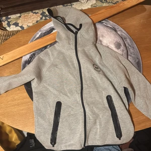 Grå hoodiejacka från WA Streetwear - Snygg grå hoodiejacka från WA Streetwear med svart tryck på bröstet. Jackan har hel dragkedja, två fickor med svarta dragkedjor och en skön huva. Perfekt för chill dagar och streetstyle. Materialet är mjukt och känns som bomull eller syntet.