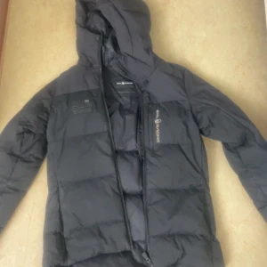 Svart pufferjacka från Sail Racing - Svart pufferjacka från Sail Racing med huva och dragkedja framtill. Jackan har en quiltad design och logotyp på bröstet. Perfekt för kalla dagar och har en sportig look som passar till streetwear. Flera praktiska fickor och snygg passform. Är som ny, skriv för fler bilder. Köptes på stadium för 3000, pris kan diskuteras 