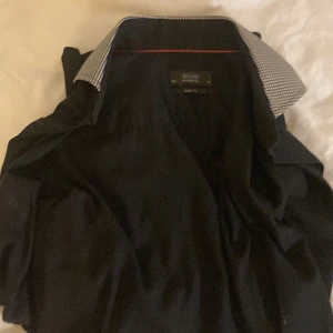 Svart Eton slim fit skjorta - Svart skjorta från Eton i slim fit med snygga rutiga detaljer på krage och manschetter. Klassisk design med knappar framtill och lång ärm. Perfekt för dig som gillar stilrena och moderna plagg med en twist.