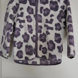 Lila teddyjacka med leopardmönster - Mysig teddyjacka från Mywear Young i storlek 110/116. Jackan är vit med stora lila leopardfläckar, har lång ärm och dragkedja framtill. Perfekt för dig som gillar djurmönster och vill ha en varm, fluffig look. Jackan har rundad nederkant och mjukt fleece-material.